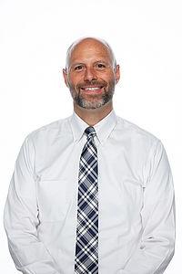 Howard Munson stands in front of a white background, he is facing forward and smiling at the camera.